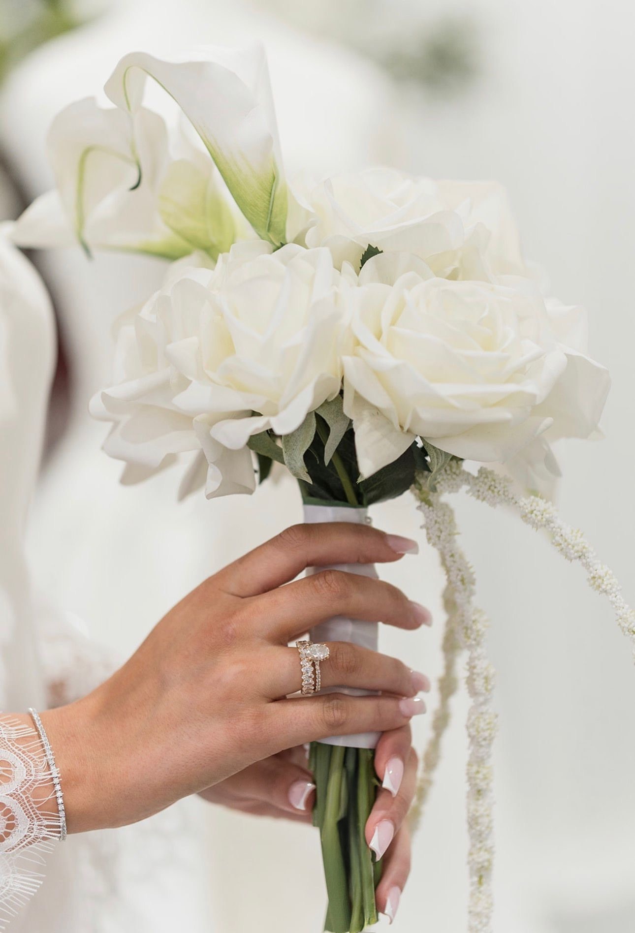 Engagement ring on a bride's hand resting on a white rose and calla lily bouquet